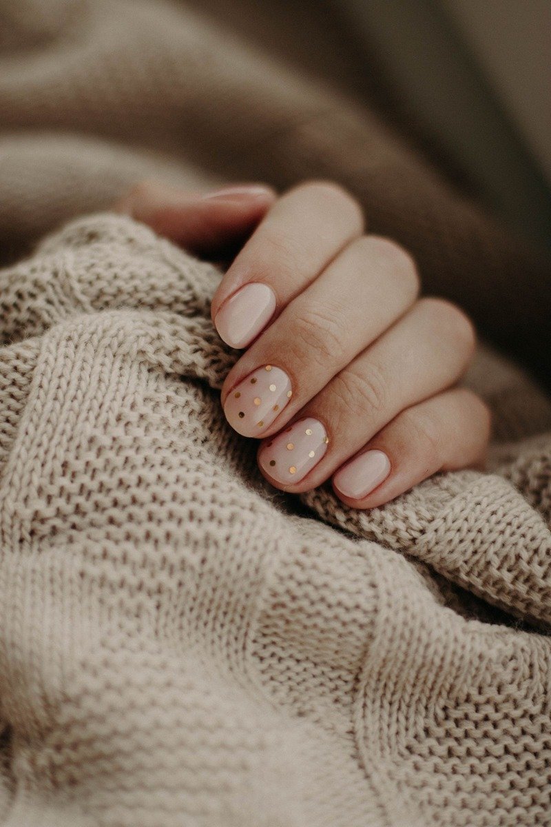 Nude almond nails with tiny gold polka dots on accent nails, hand in chunky beige knit sweater