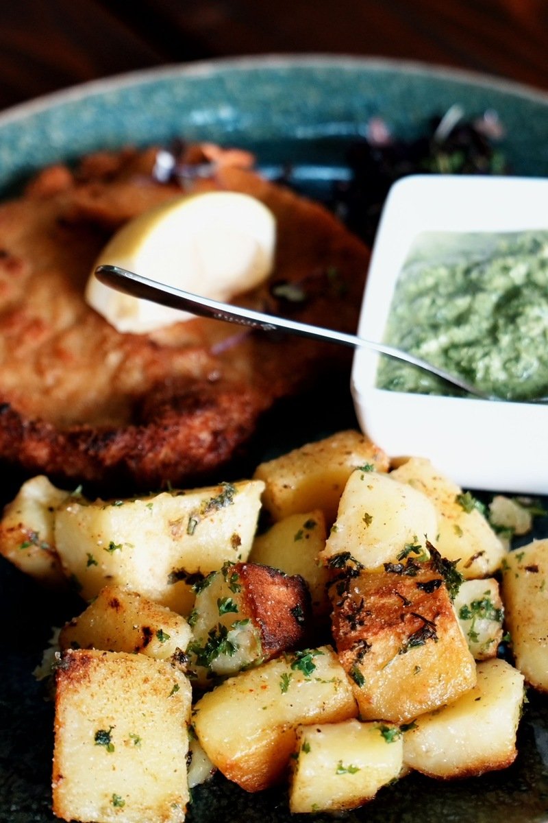 Breaded schnitzel with roasted potatoes, green sauce, and a lemon wedge on a plate
